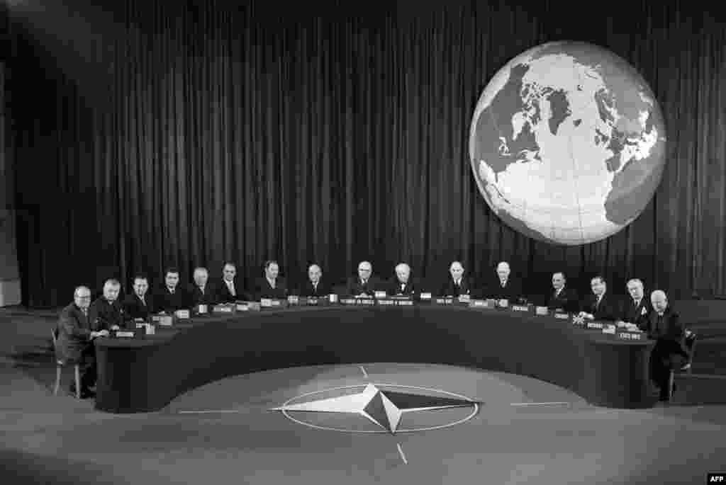 A NATO conference in Paris in 1957 behind the NATO emblem which was adopted in 1953. NATO's flag features a dark blue, representing the Atlantic Ocean, with a compass rose representing the direction toward peace, and a circle symbolizing unity. 